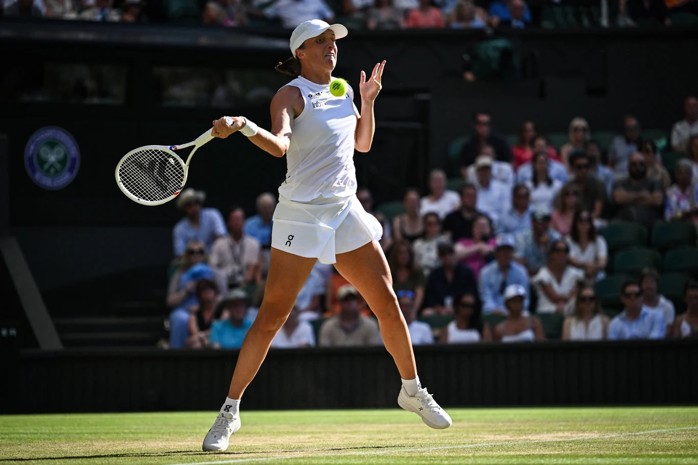 Tenisistka w białym stroju wykonuje dynamiczne uderzenie na korcie trawiastym, w tle widownia oraz elementy stadionu.