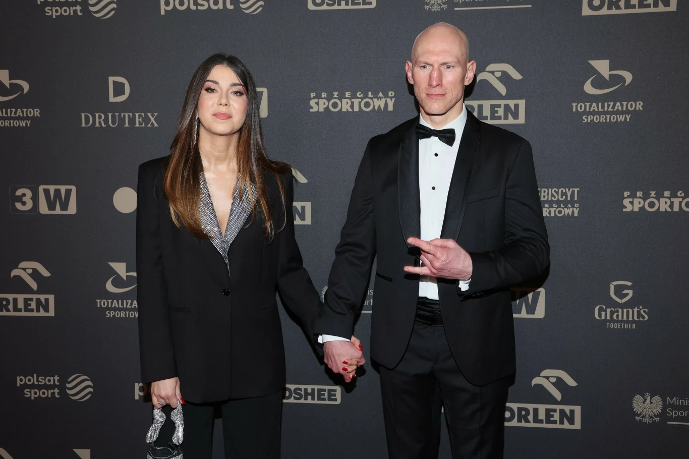 Para ubrana elegancko stoi razem na tle ściany z logotypami sponsorów, trzymając się za ręce; kobieta w ciemnym garniturze, mężczyzna w smokingu i z gestem dłoni.