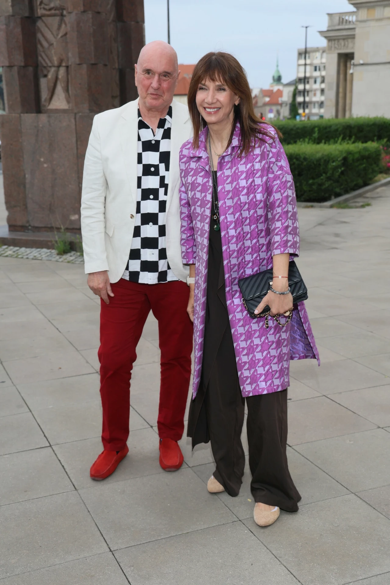 Grażyna Wolszczak i Cezary Harasimowicz zaskoczyli wszystkich oryginalną stylizacją Mężczyzna w jasnej marynarce, koszuli w czarno-białą szachownicę i czerwonych spodniach stoi obok kobiety w długim fioletowym płaszczu w geometryczny wzór, z czarną torebką w ręku. Oboje uśmiechnięci, pozują do zdjęcia na tle miejskiego krajobrazu.