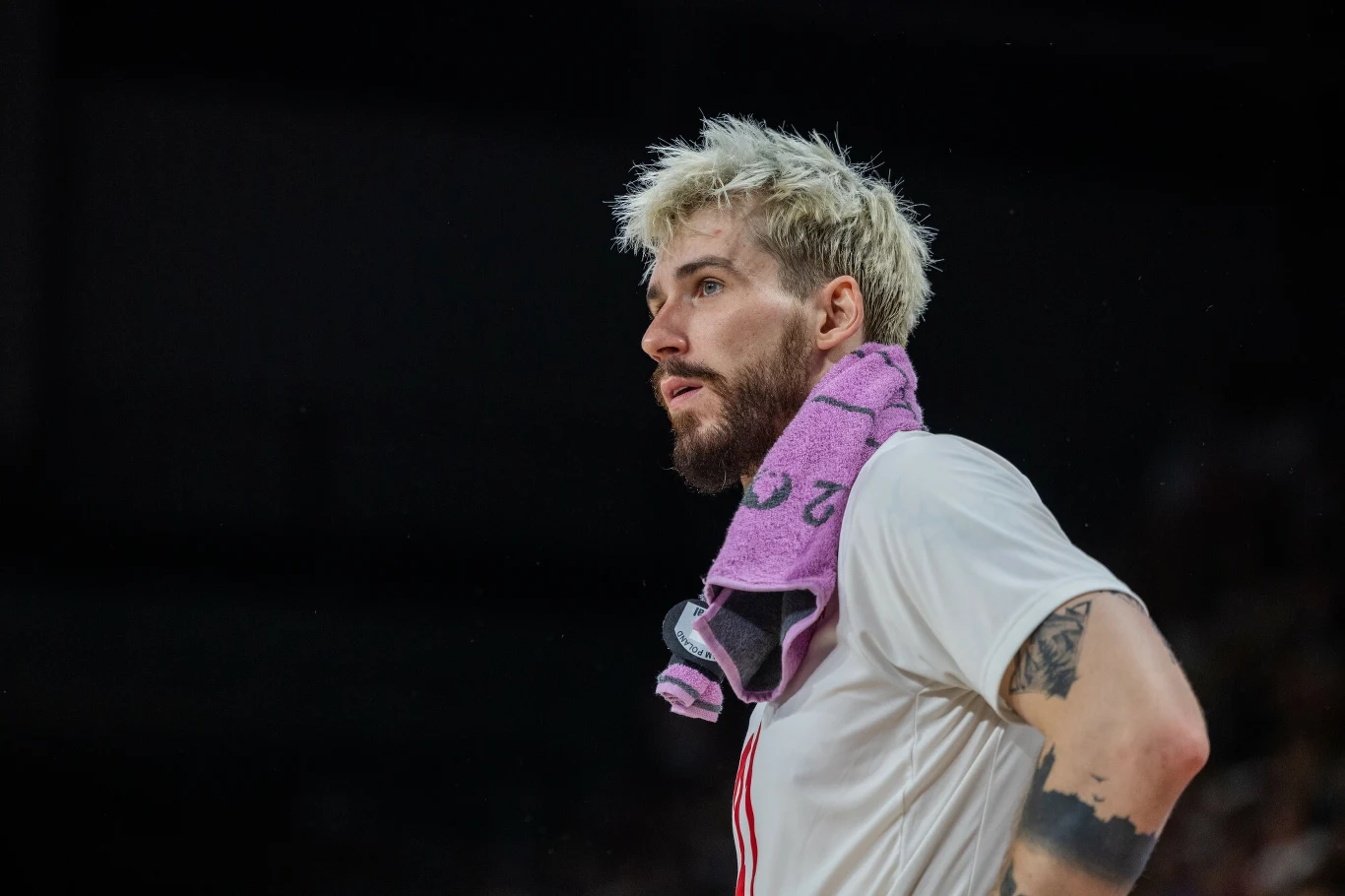 Tomasz Fornal Sportowiec z jasnymi, krótkimi włosami i zarostem w białej koszulce, z różowym ręcznikiem zawieszonym na szyi; w tle rozmyta ciemna przestrzeń.