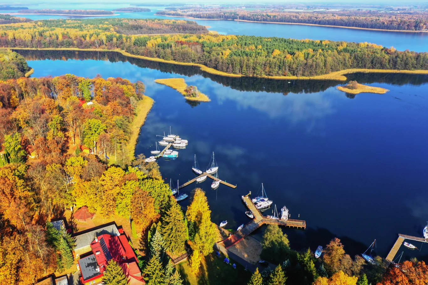 Urlop na Warmii i Mazurach? Teraz to łatwiejsze - rusza pilotażowy program dopłat w ramach bonu turystycznego! Jezioro otoczone lasami w kolorach jesieni, kilka marin z zacumowanymi jachtami oraz zabudowania rekreacyjne widoczne wśród drzew, w tle rozległe tereny leśne i dalsze brzegi zbiornika wodnego.