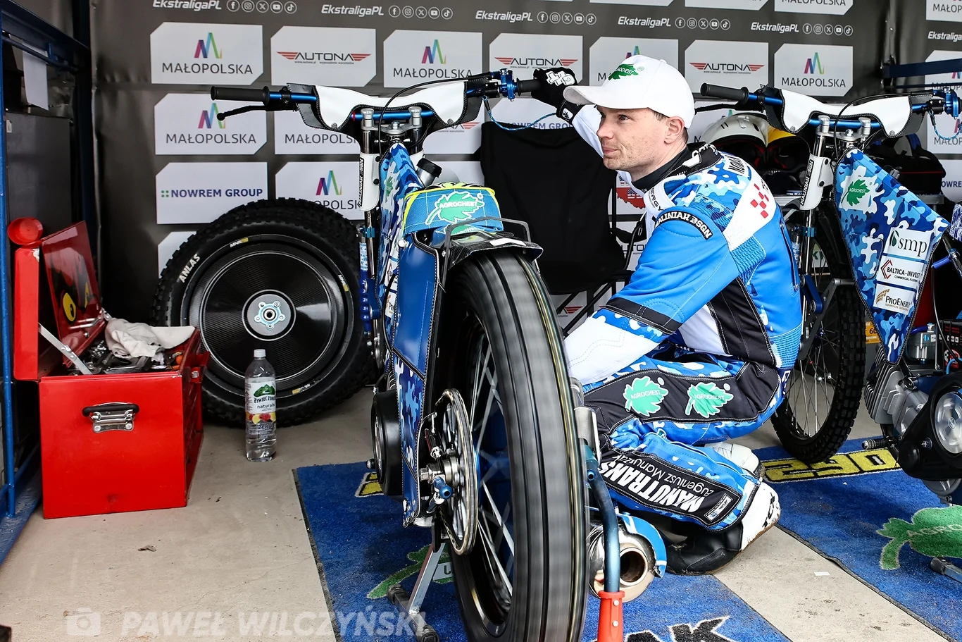 Zawodnik żużlowy ubrany w niebiesko-biały kombinezon siedzi przy motocyklu żużlowym w boksie, wokół znajdują się narzędzia, zapasowe koło i banery sponsorów.