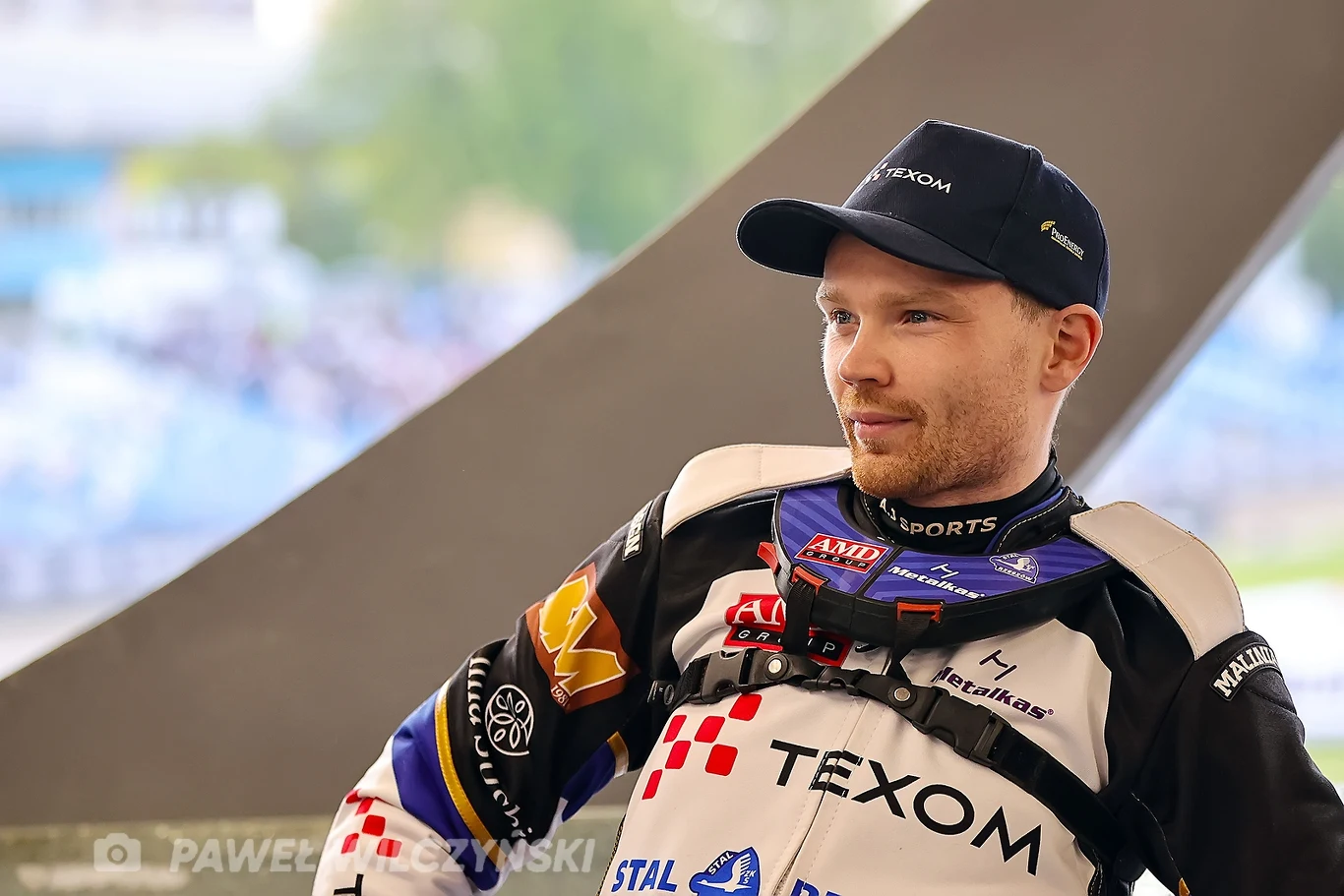 Zawodnik żużlowy w kombinezonie sportowym z licznymi logotypami sponsorów i herbem klubu, siedzący ze spokojnym wyrazem twarzy, w tle rozmyty stadion sportowy.