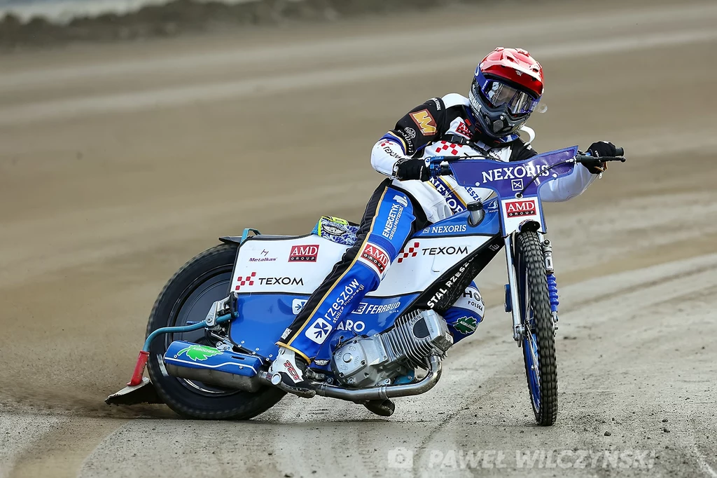Marcin Nowak Motocyklista w kombinezonie i kasku podczas dynamicznego zakrętu na torze żużlowym, motocykl pochylony, w tle rozmazana powierzchnia toru.