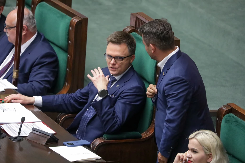 Szymon Hołownia Kilku mężczyzn w garniturach rozmawia ze sobą, siedząc w ławach sejmowych, jeden z nich odwraca się i gestykuluje podczas rozmowy, z przodu widoczna kobieta z blond włosami