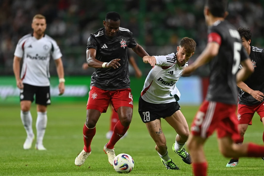 Legia Warszawa - FK Aktobe Dwóch piłkarzy walczy o piłkę na boisku podczas meczu, jeden ubrany jest w czarny strój, drugi w biały, w tle widoczni inni zawodnicy oraz trybuny stadionu.