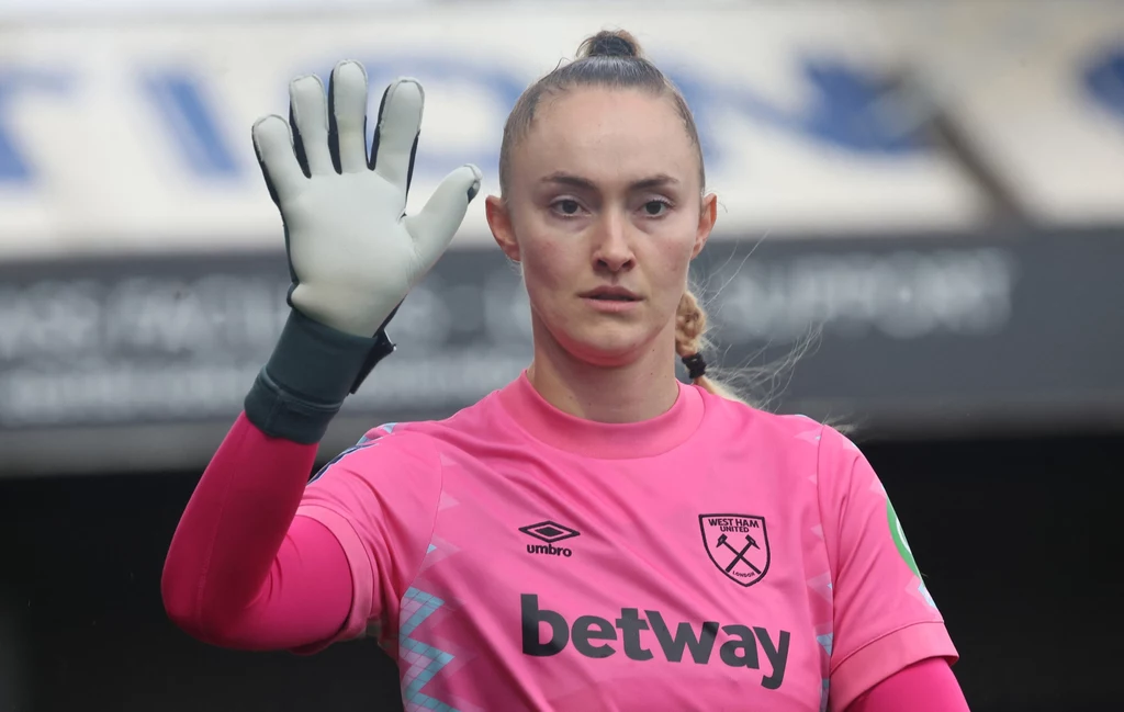 Kobieta w różowym stroju sportowym z logo klubu West Ham United i rękawicą bramkarską unosi dłoń w geście powitania lub sygnału, patrząc prosto w obiektyw.