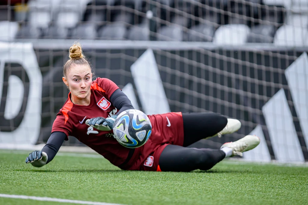 Bramkarka w czerwonym stroju sportowym dynamicznie broni piłkę podczas treningu na boisku piłkarskim, leżąc na murawie, z pełnym skupieniem na twarzy.