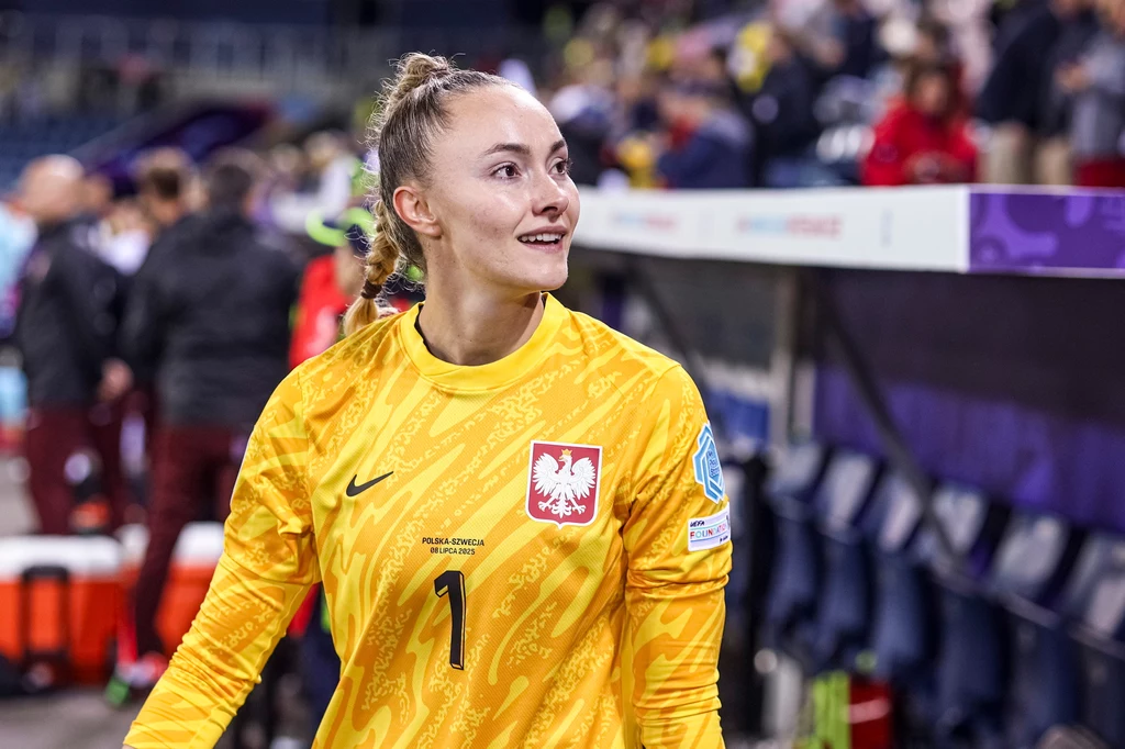 Kobieta w żółtym stroju bramkarskim z orłem na piersi stoi na stadionie, w tle widać rozmyte postacie oraz elementy trybun.