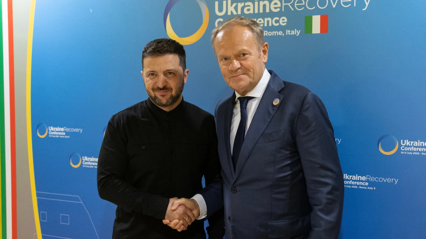 Wołodymyr Zełenski spotkał się z Donaldem Tuskiem. Padły podziękowania Dwaj mężczyźni w eleganckich ubraniach podają sobie ręce na tle niebieskiej ścianki z logo i napisem dotyczącym konferencji odbudowy Ukrainy w Rzymie, flaga Włoch po lewej stronie.