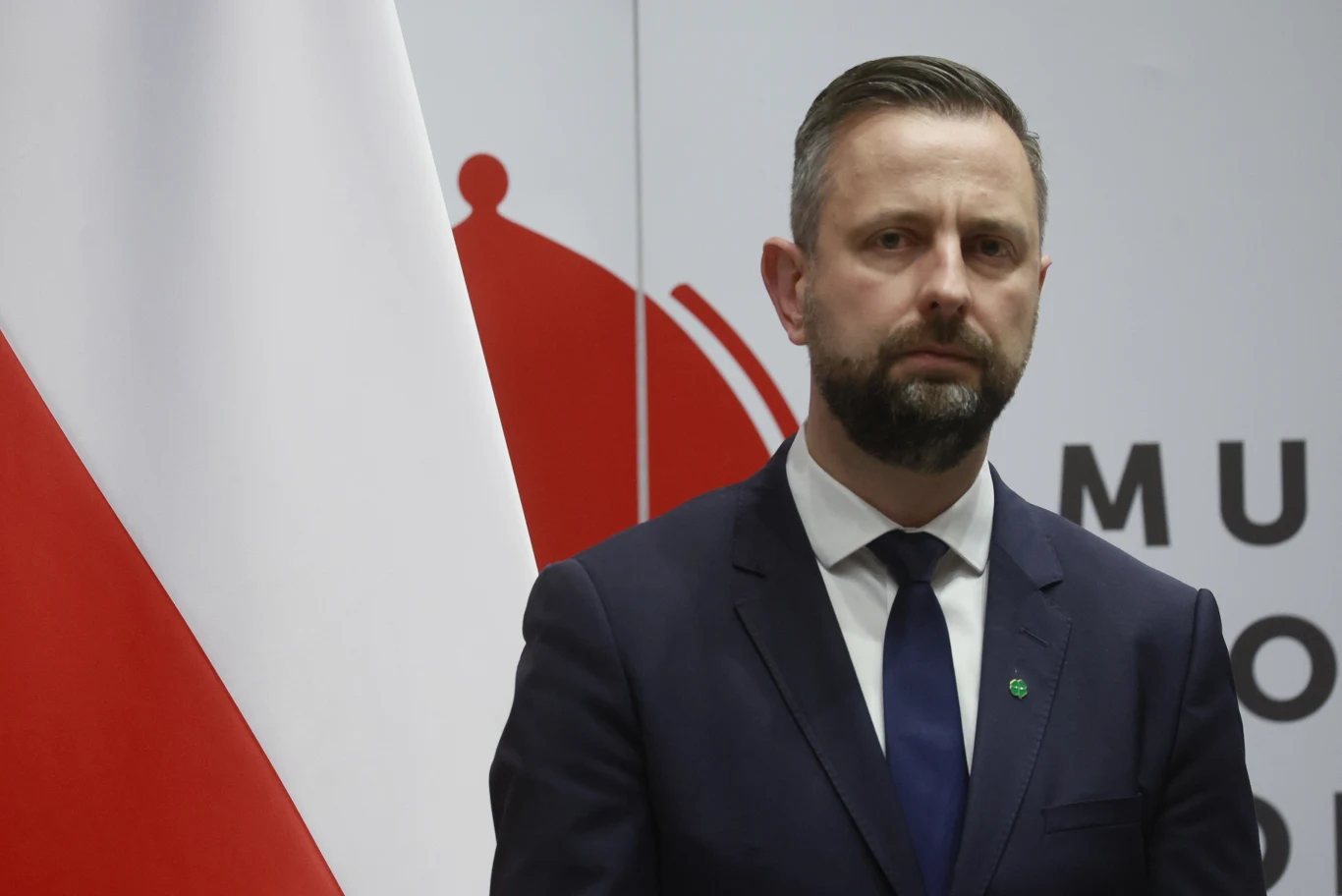 Wicepremier Władysław Kosiniak-Kamysz Mężczyzna w garniturze i krawacie stoi na tle polskiej flagi oraz fragmentu logo instytucji. Jego wyraz twarzy jest poważny i skupiony, scena ma oficjalny charakter.