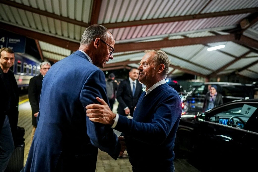 Kanclerz Niemiec Friedrich Merz i premier Polski Donald Tusk Dwóch mężczyzn w eleganckich ubraniach uściska sobie dłonie na peronie kolejowym, w tle widoczne czarne samochody i kilku innych mężczyzn. Sufit z metalowych konstrukcji oraz oświetlenie podkreślają atmosferę spotkania.