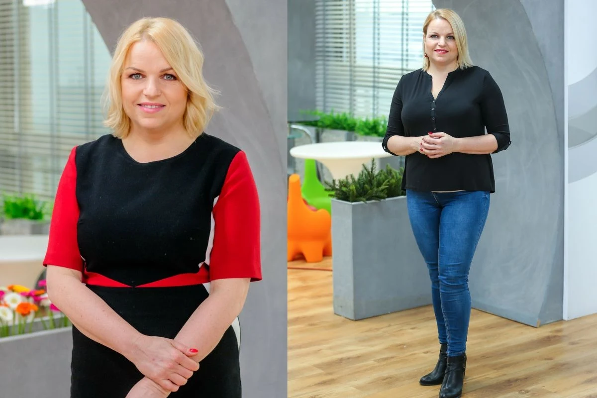 Kobieta o blond włosach pozuje w jasnym, nowoczesnym wnętrzu, częściowo wśród zieleni i kwiatów, ubrana w elegancką sukienkę z czerwonymi rękawami oraz czarne spodnie z czarną bluzką i botkami.