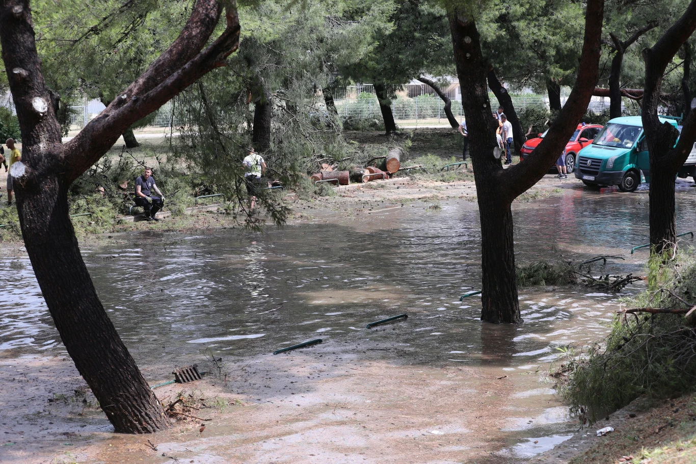 Zniszczenia w okolicy stadionu Poljud po burzy, która zdewastowała Split Podtopione drzewa w parku, woda zalewająca ziemię i fragmenty dróg, w tle zielone samochody i kilka osób obserwujących skutki powodzi.
