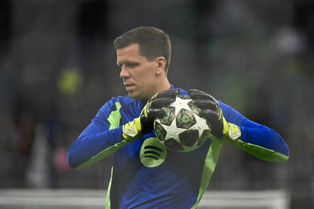 Wojciech Szczęsny Bramkarz w sportowym stroju trzymający piłkę do gry w piłkę nożną na stadionie, w tle zamazane sylwetki widzów.