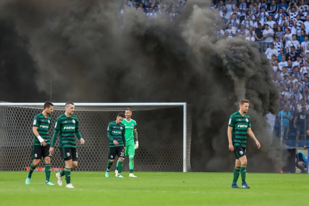 Przerwany mecz Lech Poznań - Legia Warszawa Piłkarze drużyny w zielono-czarnych strojach przemieszczają się po murawie podczas meczu, w tle widoczna ogromna chmura czarnego dymu unosząca się za bramką, a trybuny wypełnione są kibicami.