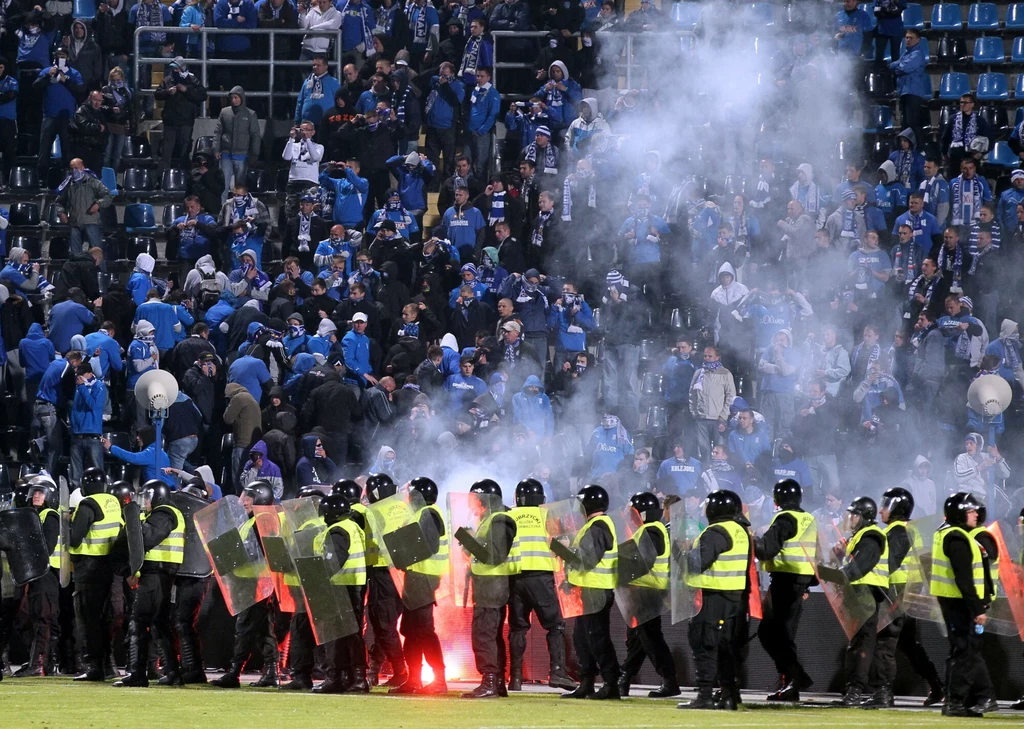 Burdy po meczu Lech Poznań - Legia Warszawa w 2011 roku Policjanci uzbrojeni w tarcze i kamizelki odblaskowe ustawieni w linii oddzielają dymiący tłum kibiców na stadionowych trybunach, wokół widoczny gęsty dym i race.
