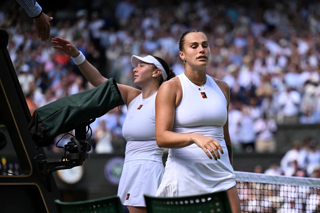 Aryna Sabalenka po porażce w półfinale Wimbledonu Dwie tenisistki idące przez kort tenisowy po zakończonym meczu, ubrane w białe stroje sportowe, jedna z nich z wyrazem zmęczenia lub rozczarowania na twarzy, w tle rozmyta publiczność.