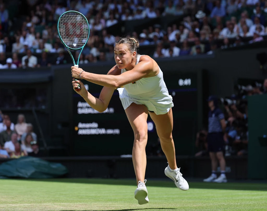 Aryna Sabalenka, Wimbledon 2025 Tenisistka w białym stroju wykonuje dynamiczny ruch podczas meczu na trawiastym korcie, w tle widownia i sędzia oraz tablica wyników.