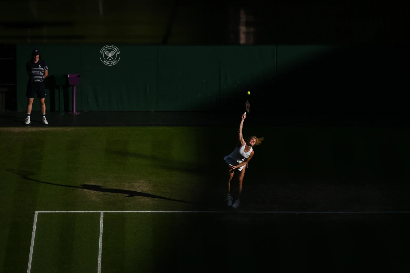 Katerina Siniakova podczas finału Wimbledonu w grze mieszanej Zawodniczka na trawiastym korcie tenisowym w trakcie serwisu, piłka tenisowa znajduje się wysoko nad jej głową, tło w znacznej części spowite cieniem, z boku stoi sędzia liniowy.