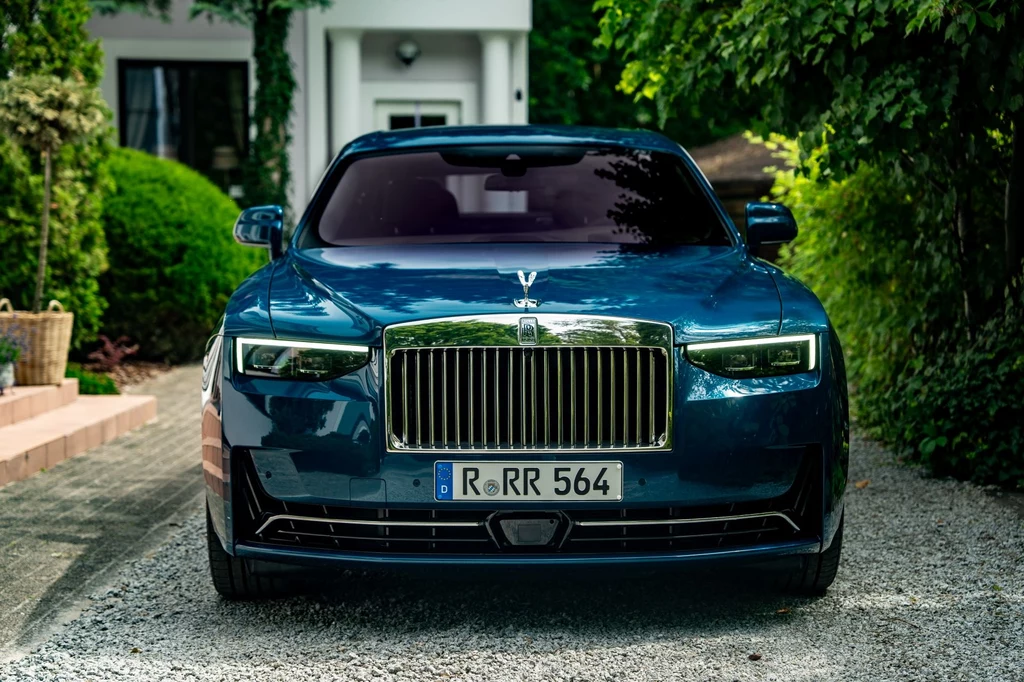 Po modernizacji z 2024 roku Rolls-Royce Ghost wyróżnia się zmienionymi światłami i przeprojektowanym zderzakiem, mającym bardziej podkreślać osłonę chłodnicy "Pantheon". Luksusowy ciemnoniebieski samochód marki Rolls-Royce zaparkowany na żwirowej alei, w otoczeniu zieleni i nowoczesnego budynku w tle. Imponujący grill, reflektory i tablica rejestracyjna podkreślają prestiż pojazdu.