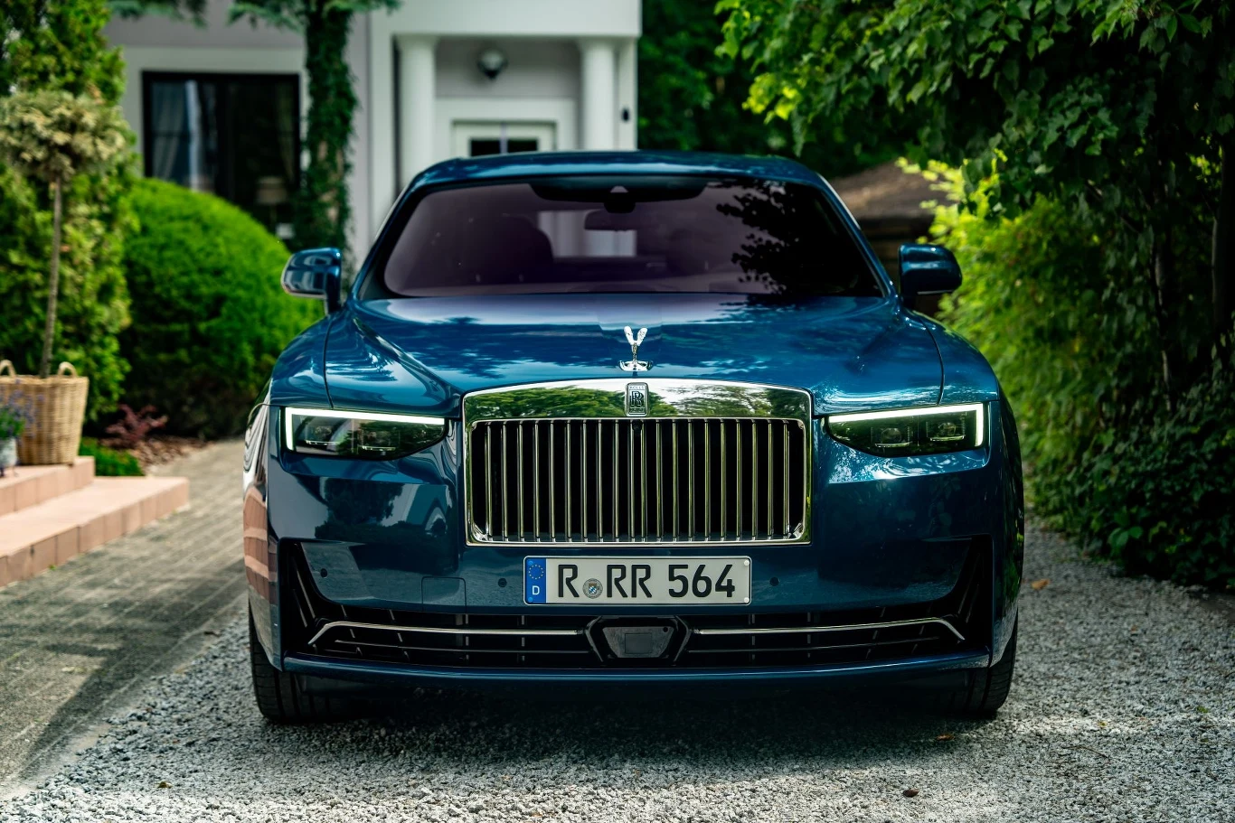 Luksusowy ciemnoniebieski samochód marki Rolls-Royce zaparkowany na żwirowej alei, w otoczeniu zieleni i nowoczesnego budynku w tle. Imponujący grill, reflektory i tablica rejestracyjna podkreślają prestiż pojazdu.