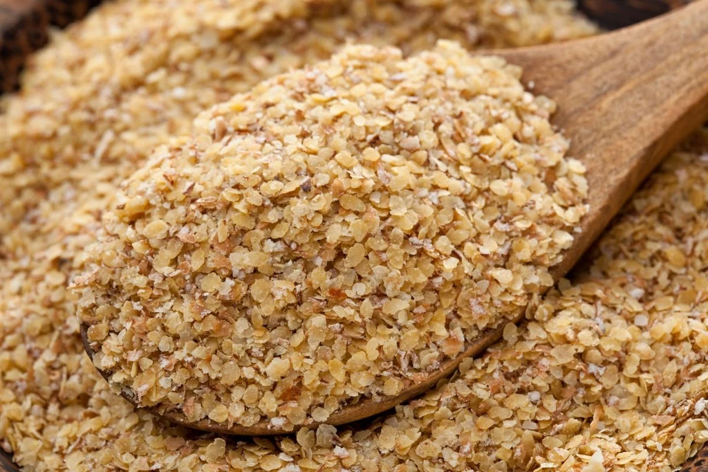 Drewniana łyżka wypełniona bulgurem, drobnoziarnistą pszenicą o jasnobrązowej barwie, ziarna rozsypane częściowo wokół łyżki tworząc naturalny, spożywczy motyw.