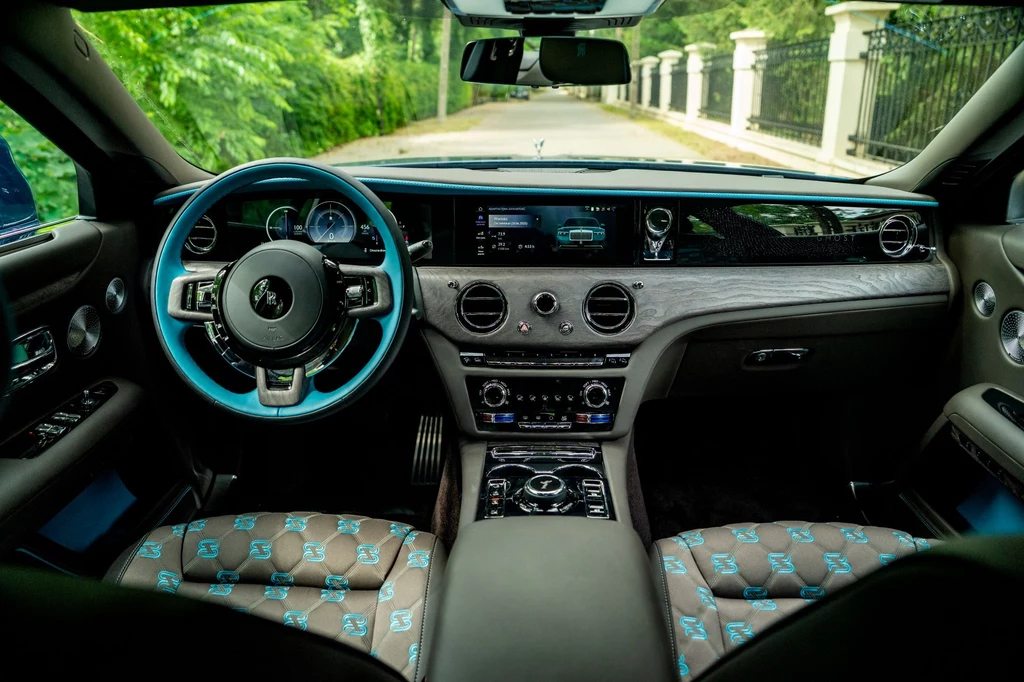 Wnętrze Rolls-Royce'a Ghosta sprawnie łączy teraźniejszość (ekrany) z przeszłością (fizyczne przełączniki, pokrętła i dźwignie). Luksusowe wnętrze samochodu z nowoczesną deską rozdzielczą, dwukolorową kierownicą i wysokiej klasy skórzanymi siedzeniami ozdobionymi monogramem, widok zza kierownicy na zieloną alejkę przed pojazdem.