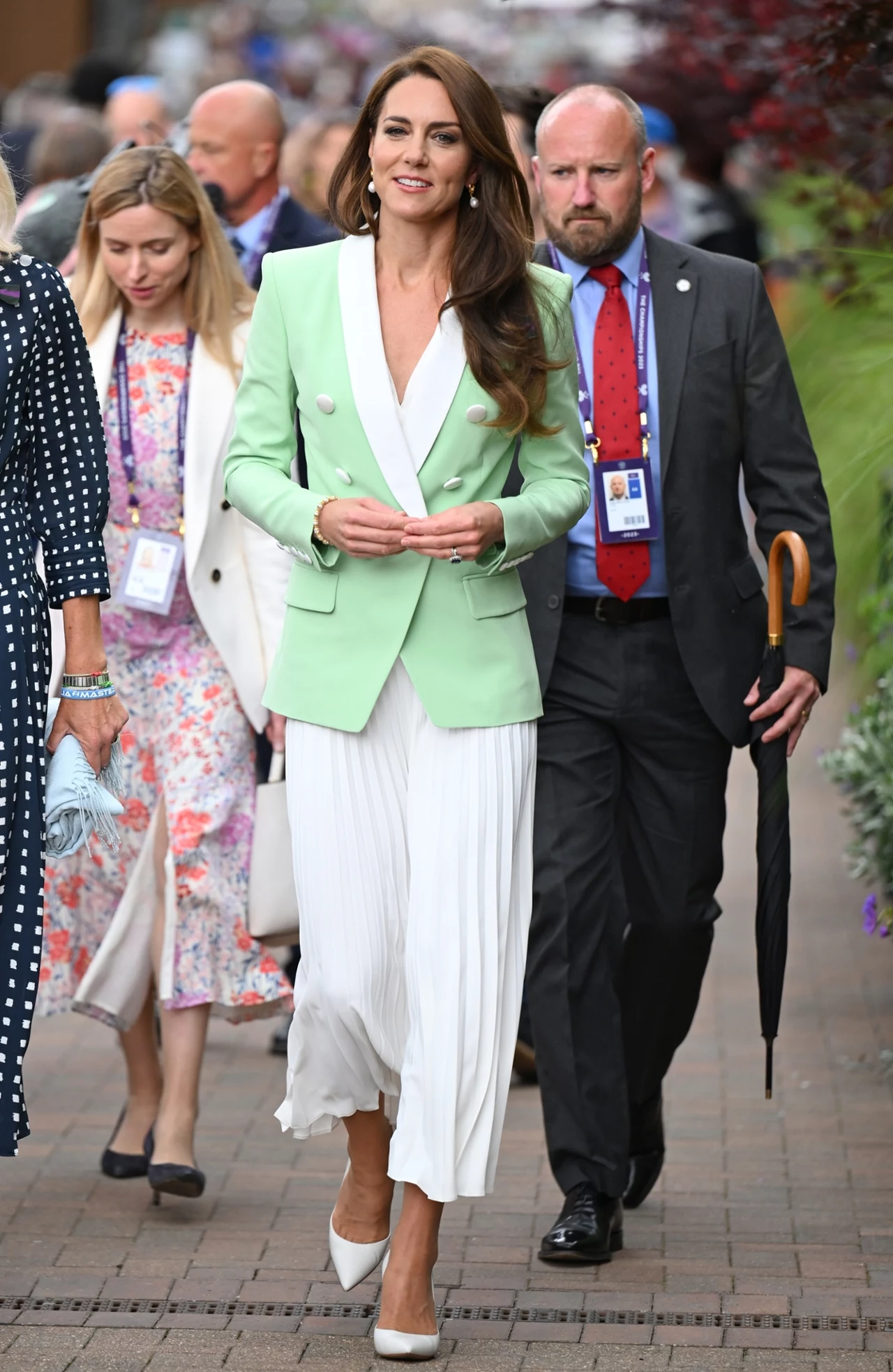 Elegancko ubrana kobieta w jasnozielonej marynarce i białej, plisowanej spódnicy idzie chodnikiem, otoczona przez kilka osób, w tym mężczyznę z plakietką i parasolem oraz kobietę w długiej sukience w kwiaty.