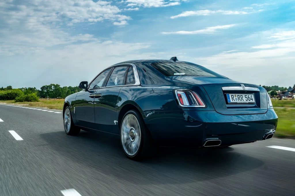 Rolls-Royce Ghost rozpędza się do 100 km/h w 4,8 sekundy. Warto podkreślić, że to auto ważące blisko 2,5 tony. Luksusowy czarny sedan jadący dynamicznie po asfaltowej drodze na tle letniego krajobrazu i błękitnego nieba, reflektory i detale nadwozia podkreślają elegancję i prestiż samochodu.