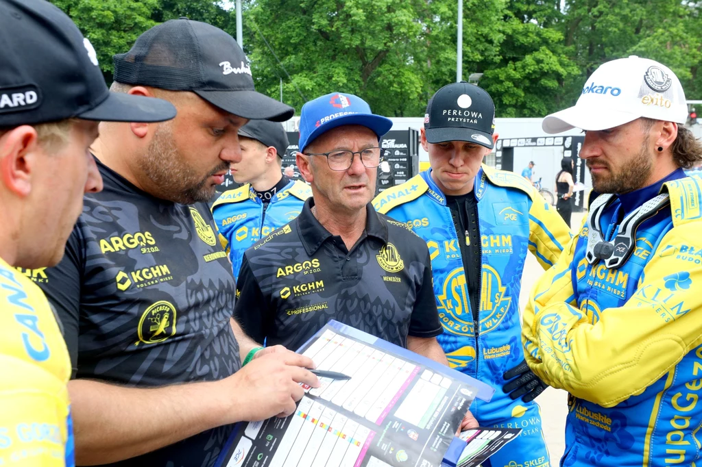 Stal Gorzów podczas odprawy meczowej Grupa mężczyzn ubranych w sportowe stroje żużlowe podczas analizy strategii na bieżącym spotkaniu, trzymają w rękach kartkę z rozpisanymi planami, skoncentrowani w rozmowie, w tle zielone drzewa.