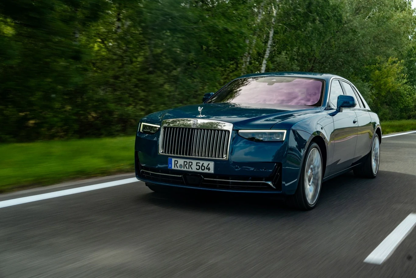 Luksusowy samochód marki Rolls-Royce Phantom w ciemnoniebieskim kolorze jedzie po asfaltowej drodze otoczonej zielenią, w tle drzewa i zarośla, auto zajmuje centralną część kadru.