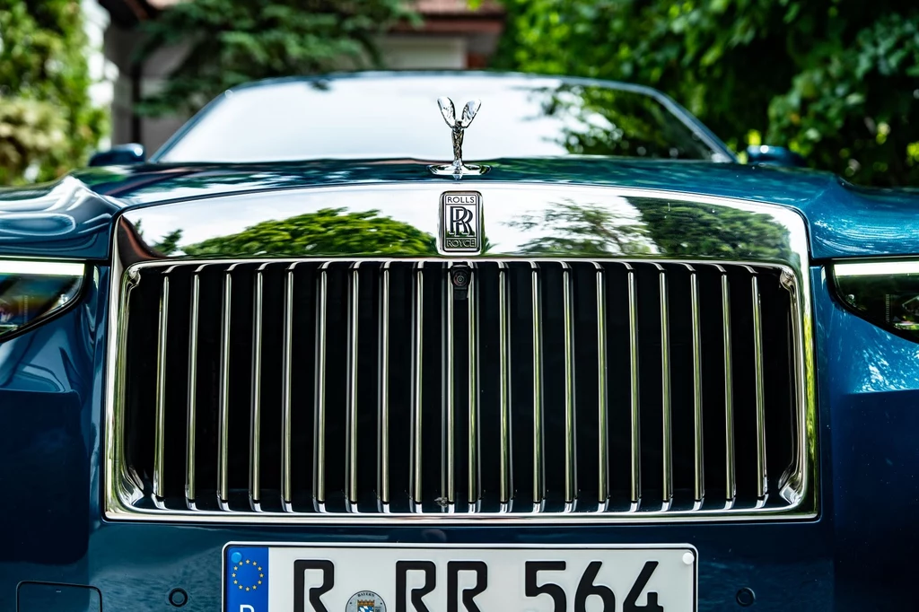 Słynna osłona chłodnicy Rolls-Royce'a, Pantheon, jest podświetlana. Chromowana atrapa chłodnicy oraz charakterystyczna figurka Spirit of Ecstasy na masce luksusowego samochodu Rolls-Royce, z widoczną tablicą rejestracyjną i błyszczącym, niebieskim lakierem, otoczony zielenią w tle.