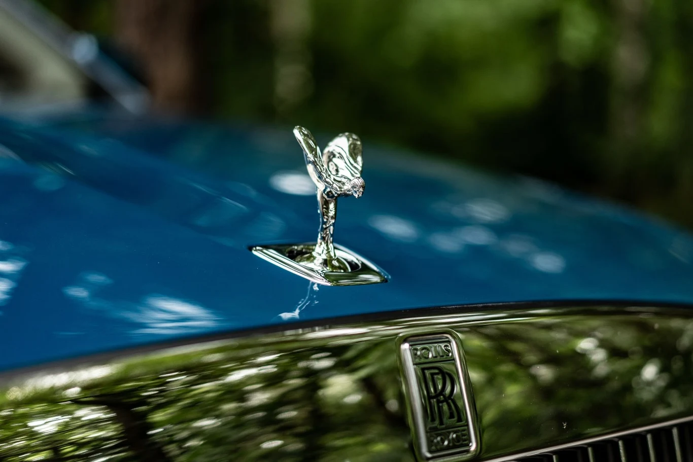 Figurka "Spirit of Ecstasy" zdobiąca przód Rolls-Royce. Maskotka Spirit of Ecstasy umieszczona na masce niebieskiego samochodu marki Rolls-Royce z wyraźnym logo producenta na tle rozmytych drzew.