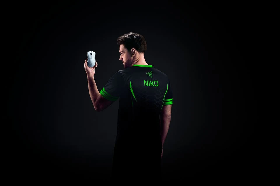 Mężczyzna ubrany w czarną koszulkę z zielonymi akcentami oraz napisem 'NIKO' na plecach, trzyma w lewej dłoni biały sprzęt elektroniczny na tle ciemnego, minimalistycznego tła.