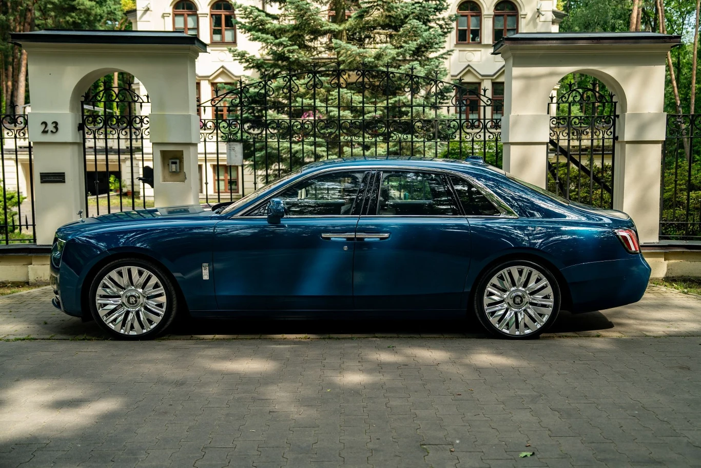 Luksusowa niebieska limuzyna zaparkowana na ulicy przed eleganckim ogrodzeniem z kutego żelaza i reprezentacyjną kamienicą w tle, otoczoną zielenią i wysokimi drzewami.