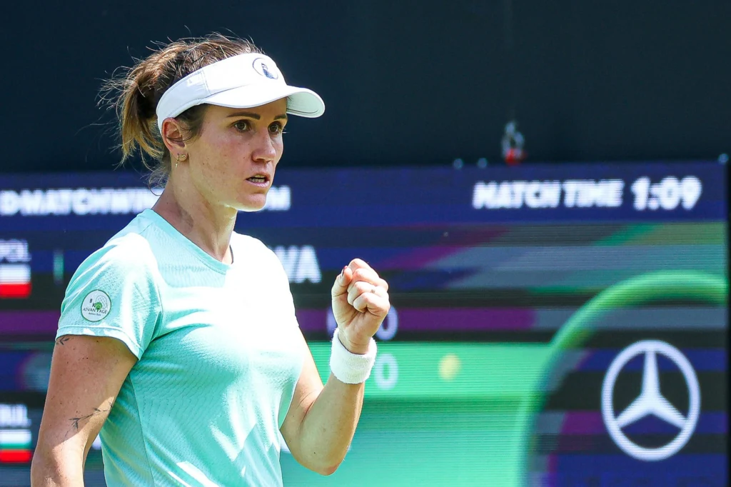 Tenisistka na korcie w dynamicznej pozie, zaciskająca pięść w geście triumfu, w tle tablica wyników i logo sponsora motoryzacyjnego.