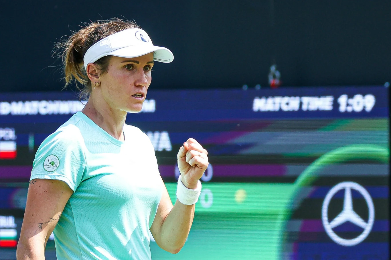 Tenisistka na korcie w dynamicznej pozie, zaciskająca pięść w geście triumfu, w tle tablica wyników i logo sponsora motoryzacyjnego.