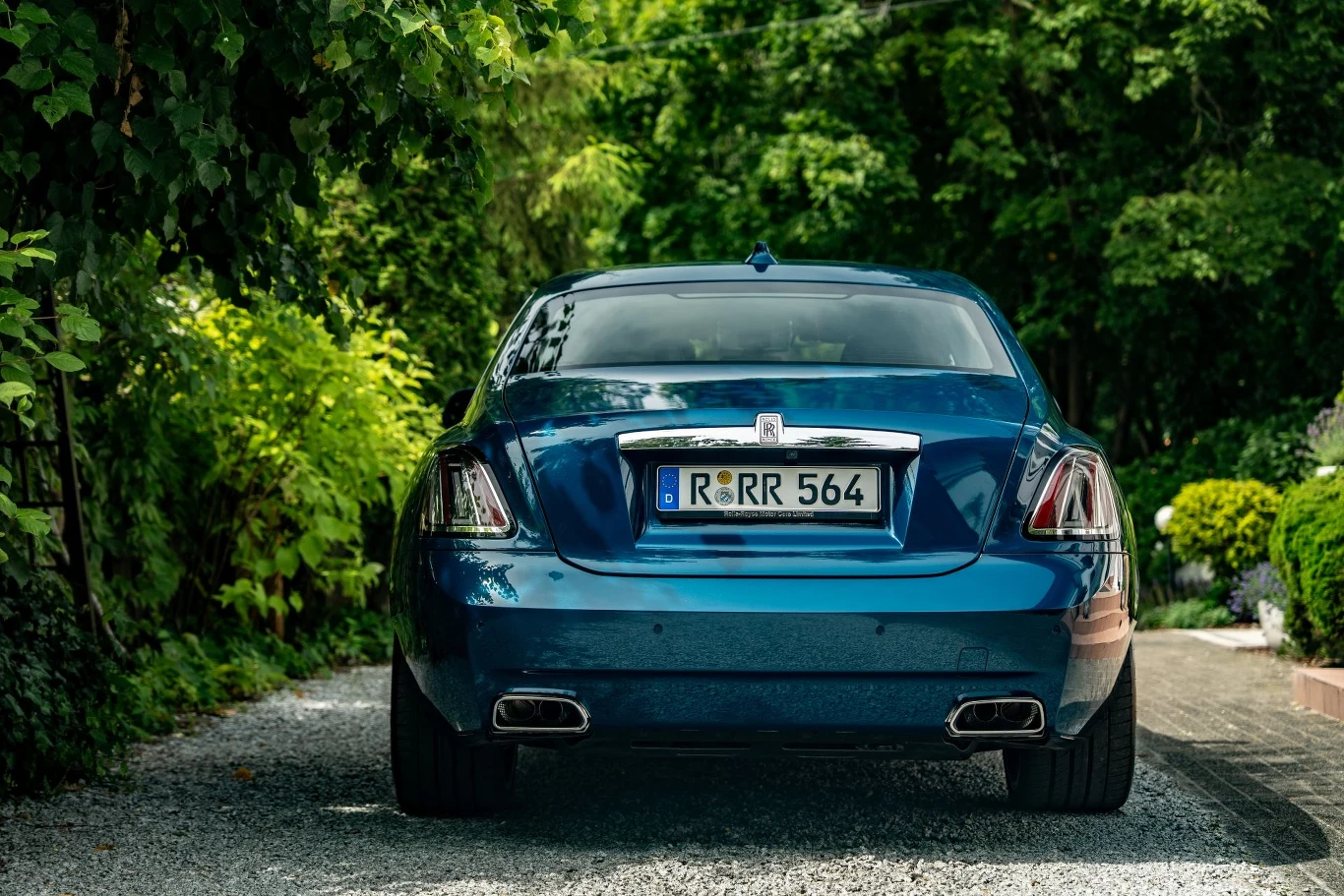 Tył luksusowego, niebieskiego samochodu marki Rolls-Royce zaparkowanego na żwirowej alejce otoczonej gęstą zielenią i ogrodową roślinnością.