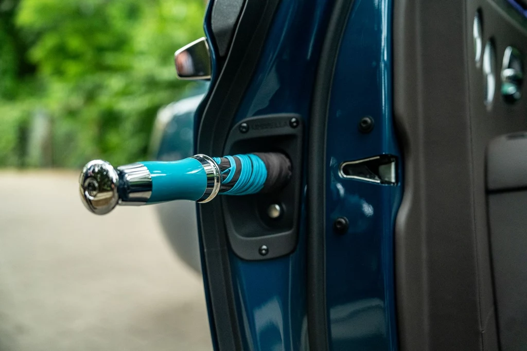 Uwagę zwracają również takie detale, jak znany z innych modeli Rolls-Royce'a parasol ukryty w drzwiach. Kolorowa parasolka schowana wewnątrz drzwi samochodu, z widocznym połyskującym uchwytem i detalami, w tle rozmyta zieleń.