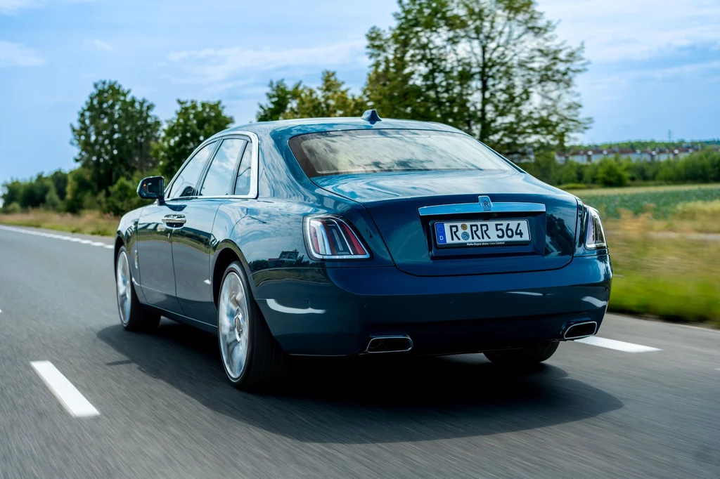 Dzięki mocy 571 KM i 850 Nm ważący blisko 2,5 tony Ghost osiąga 100 km/h w 4,8 sekundy. Prędkość maksymalna to 250 km/h. Luksusowy sedan w kolorze granatowym jedzie drogą przez zielony, wiejski krajobraz, po lewej stronie widoczne drzewa i krzewy, na tablicy rejestracyjnej widoczny numer R RR 564.