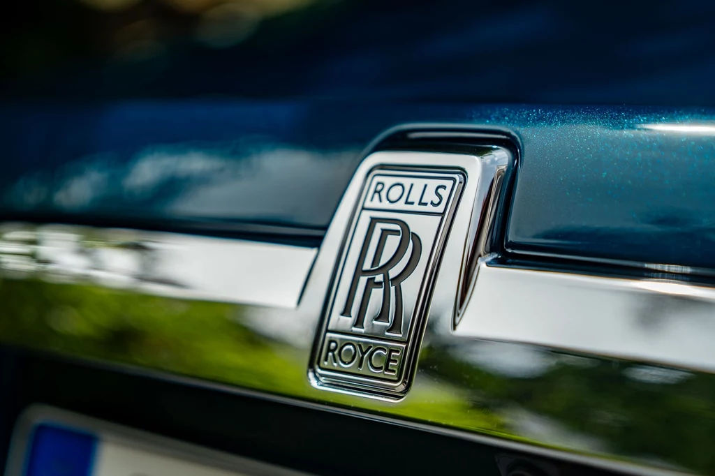 Ceny Rolls-Royce'a Ghosta w podstawowej konfiguracji startują od 235 tys. euro netto (ok. 1,2 mln zł). Chrome emblemat Rolls-Royce na błyszczącym, ciemnoniebieskim nadwoziu samochodu, wyraźne logo firmy osadzone centralnie na tle refleksów światła.