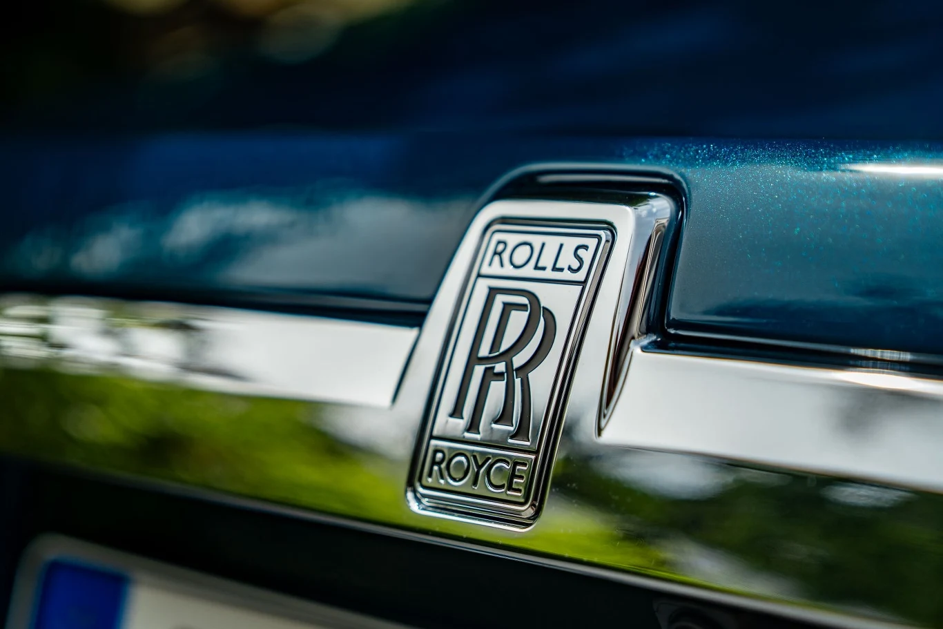 Chrome emblemat Rolls-Royce na błyszczącym, ciemnoniebieskim nadwoziu samochodu, wyraźne logo firmy osadzone centralnie na tle refleksów światła.