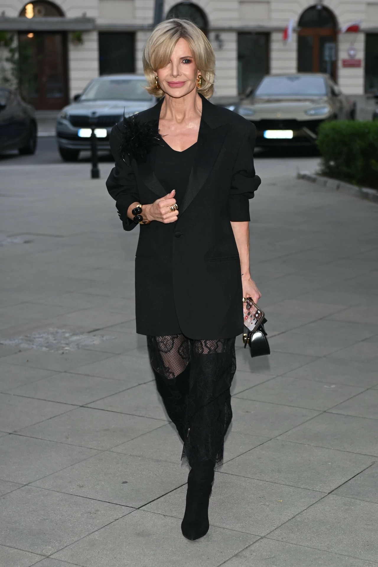 Joanna Racewicz zachwyciła w czarnej stylizacji Elegancka kobieta w czarnym żakiecie, koronkowych spodniach i czarnych butach na obcasie idzie po miejskim chodniku; trzyma w ręku ozdobną torebkę i uśmiecha się do obiektywu.