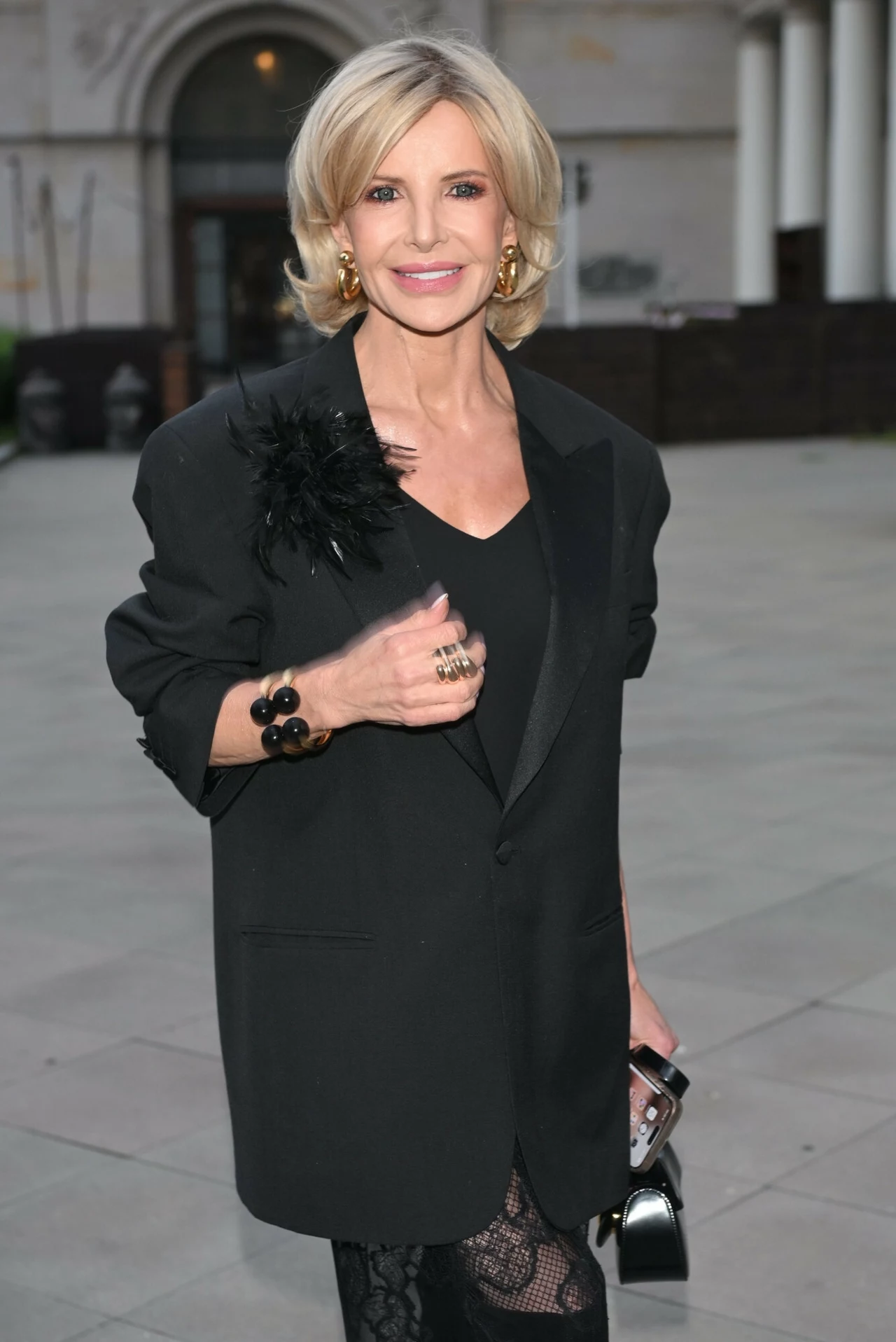 Joanna Racewicz kocha modę z wzajemnością Kobieta w średnim wieku w eleganckim czarnym żakiecie z ozdobą na ramieniu i czarną sukienką z koronkowymi wstawkami, trzyma małą torebkę, stoi na tle miejskiego placu z klasyczną architekturą w tle.