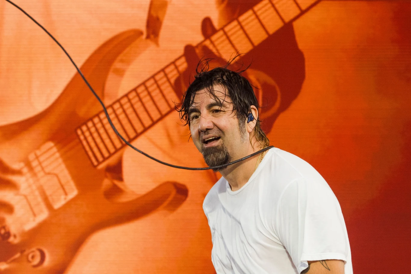 Chino Moreno (Deftones) w akcji Mężczyzna z mokrymi włosami, ubrany w biały T-shirt, stoi na scenie z mikrofonem, na tle dużej projekcji gitary elektrycznej.