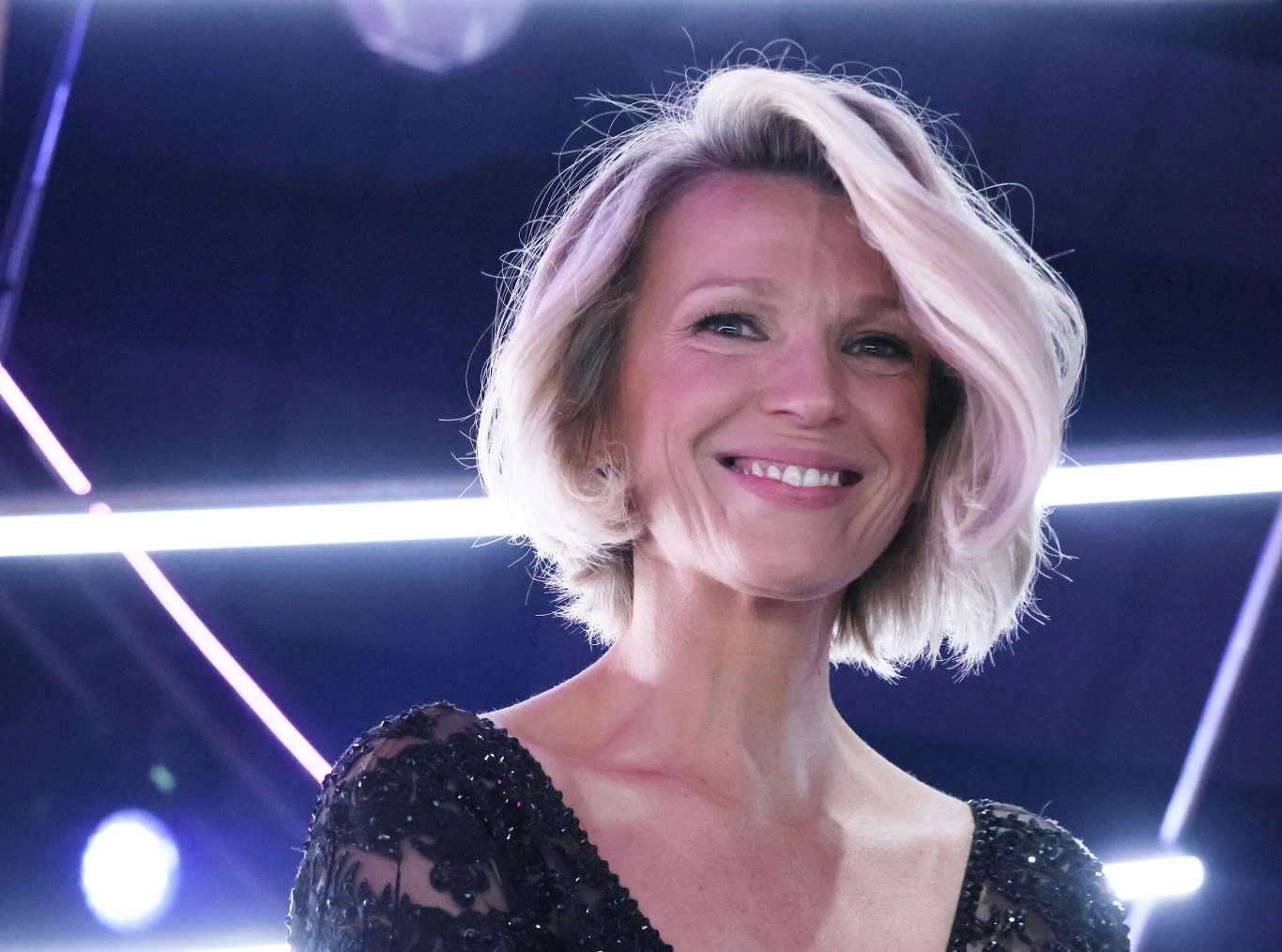 Najmodniejsza fryzura lato 2025 Uśmiechnięta kobieta z krótkimi, blond włosami, ubrana w czarną, elegancką suknię z koronkowymi rękawami, na tle jasnych, neonowych świateł.