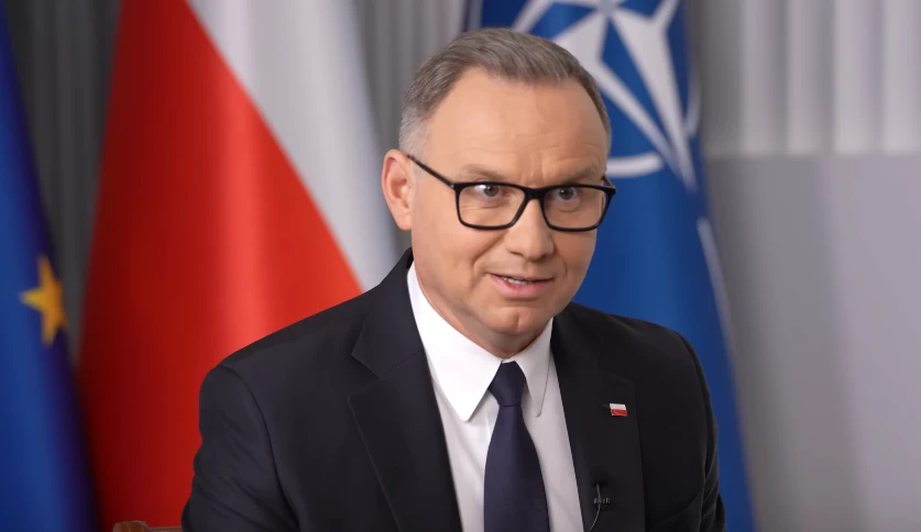 Prezydent Andrzej Duda podczas wywiadu na kanale Otwarta Konserwa Mężczyzna w garniturze z przypiętą polską flagą na klapie marynarki, noszący okulary, siedzi na tle flag Polski, Unii Europejskiej i NATO.