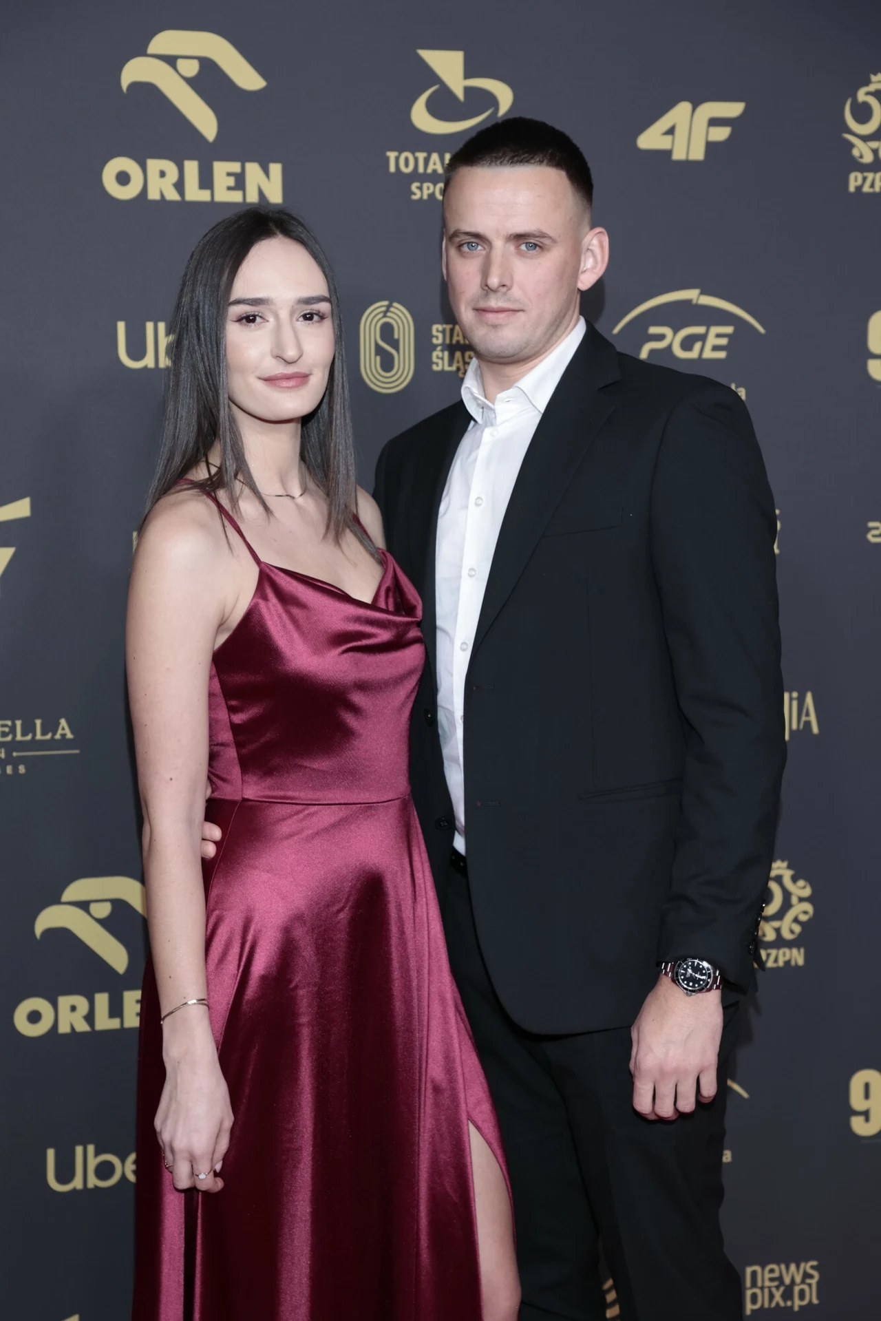 Martyna Swatowska-Wenglarczyk i Jan Wenglarczyk Elegancko ubrana para pozuje wspólnie na tle ściany z logotypami znanych marek i sponsorów, kobieta ma na sobie bordową, satynową sukienkę na ramiączkach, a mężczyzna klasyczny czarny garnitur z białą koszulą.