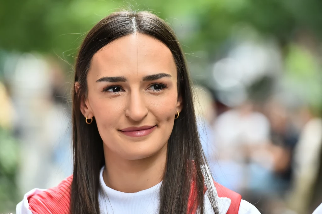 Martyna Swatowska-Wenglarczyk Młoda kobieta o długich brązowych włosach, uśmiechnięta, ubrana w sportową bluzę w kolorze białym z czerwonym akcentem, na tle rozmytego, zielonego otoczenia.